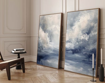 May include: Two abstract paintings in gold frames, depicting a blue and white cloudy sky over a body of water. The paintings are hanging on a white wall in a room with hardwood floors.