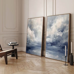 May include: Two abstract paintings in gold frames, depicting a blue and white cloudy sky over a body of water. The paintings are hanging on a white wall in a room with hardwood floors.