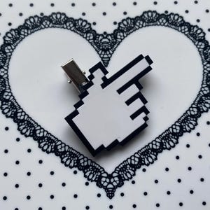 May include: A white and black pixelated cursor hair clip. The clip is in the shape of a computer cursor, with a black outline and a white fill. The clip has a silver metal clasp.