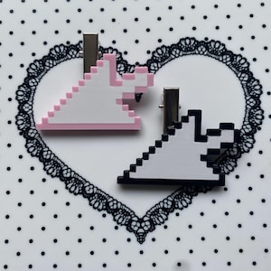 May include: Two pixelated angel-shaped hair clips. One clip is white with pink accents, the other is white with black accents. Both clips have silver metal clasps. The background is white with black polka dots and a black lace heart.