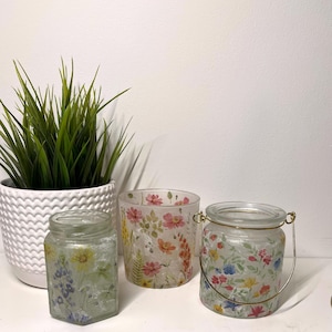 Op de afbeelding: Drie glazen kaarshouders met bloemenmotieven. De kaarshouders zijn van helder glas met een matglazen afwerking. De bloemenmotieven zijn in tinten roze, blauw en geel. De kaarshouders staan op een wit oppervlak.