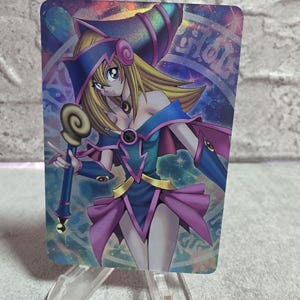 May include: A rectangular trading card featuring a holographic illustration of a blonde character wearing a blue and purple wizard hat and outfit. The character holds a gold-topped staff, set against a vibrant, swirling cosmic background.