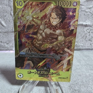 May include: A gold-foiled One Piece trading card featuring Portgas D. Ace with a 10 cost and 10000 power. The card displays Japanese text, a character illustration of a smiling figure with dark hair, and vibrant orange and yellow energy effects.