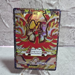 Könnte beinhalten: Eine holografische Ho-Oh EX Sammelkarte mit einer Illustration eines roten, gelben und goldenen Phönix. Die Karte zeigt 150 KP, die Attacke Phönix-Turbo und Text zu den Spielmechaniken. Der Rand hat ein dunkles, wirbelndes Musterdesign.