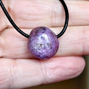 Könnte beinhalten: Ein polierter, ovaler, violetter Edelsteinanhänger an einer schwarzen Kordel. Der Stein hat ein marmoriertes Aussehen mit helleren und dunkleren Lilatönen und einigen braunen Einschlüssen. Die Kordel ist dünn und scheint aus Leder oder einem ähnlichen Material zu sein.
