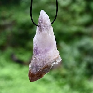 May include: A natural amethyst pendant hanging from a black cord. The crystal displays a colour gradient, ranging from a deep brown at the base to a translucent purple and white at the top. The pendant has an irregular shape, with a natural, unpolished appearance.