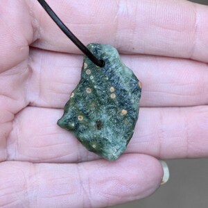 以下が含まれることがあります： 濃い茶色の革紐が付いた、緑と茶色の石のペンダント。石はざらざらとした不規則な形で、小さな丸い茶色の斑点があります。ペンダントは手で持たれています。