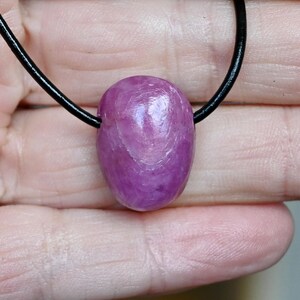 May include: A polished, oval-shaped, purple gemstone pendant on a black leather cord. The gemstone has a smooth, reflective surface with subtle internal patterns. The necklace is a simple, elegant piece of jewellery.