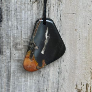 Op de afbeelding: Een gepolijste, driehoekige hanger van donkergrijze en zwarte steen met een gladde afwerking. De hanger heeft natuurlijke insluitingen van gebrand oranje en metaalachtige zilverkleurige aders. Het hangt aan een dun, zwart leren koord.