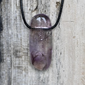 Könnte beinhalten: Ein durchscheinender, violetter Amethyst-Anhänger mit einer schwarzen Kordel. Der polierte Stein hat eine abgerundete Oberseite und einen zylindrischen Körper. Der Anhänger hängt vor einem verwitterten, grauen Holzgrund.