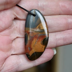 Op de afbeelding: Een gepolijste, druppelvormige stenen hanger met een glad, glanzend oppervlak met wervelende patronen van donkergrijs, dieporanje en goudbruin. De hanger hangt aan een dun zwart koord en toont natuurlijke minerale aders.