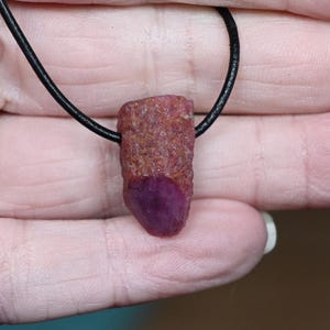 May include: A natural ruby pendant on a black cord. The ruby has a deep red-purple hue and a rough, textured appearance. The pendant hangs from a thin, black cord, forming a simple, natural necklace.