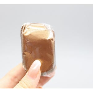 May include: A rectangular, gold-colored item wrapped in clear plastic. The item is held by a person's hand, with the focus on the wrapped product. The background is a plain, light gray.