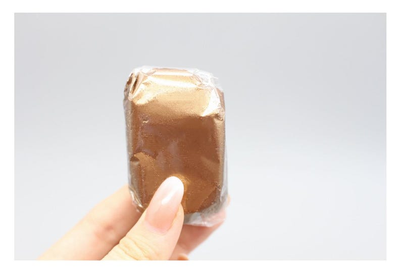 May include: A rectangular, gold-colored item wrapped in clear plastic. The item is held by a person's hand, with the focus on the wrapped product. The background is a plain, light gray.