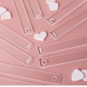 May include: Clear acrylic craft tools in various shapes and sizes, including rectangles, squares, and hearts. Measurements are labeled in millimetres, such as 3MM, 4MM, 5MM, and 6MM. The tools are arranged on a pink surface.
