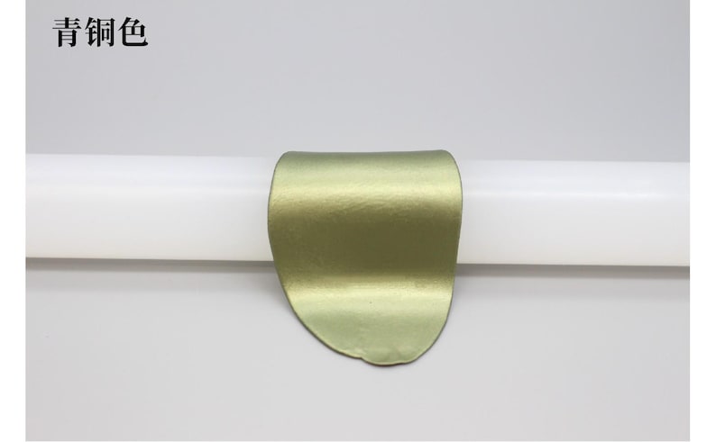 May include: A metallic, olive-green, curved object is centered on a white cylindrical form. The object has a textured surface and a rounded shape. The Chinese characters "青铜色" are in the upper left corner.