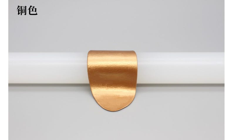 May include: A copper-colored metal clip, curved at the top and rounded at the bottom, is attached to a white cylindrical object. The clip has a textured surface. The Chinese characters "铜色" are in the top left corner.