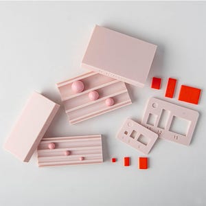 Puede incluir: Colección de formas geométricas rosas y naranjas. El conjunto incluye bloques rectangulares, bandejas a rayas con esferas rosas y un calibrador rosa con recortes numerados. El texto "10-12-15 mm" es visible.