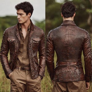 Puede incluir: Chaqueta de cuero marrón con textura de piel de cocodrilo. La chaqueta tiene dos bolsillos en el pecho, dos bolsillos laterales y un cinturón. El modelo lleva una camisa y pantalones marrones. El fondo es una escena exterior borrosa.