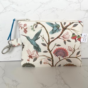May include: A small white zippered pouch with a floral print featuring hummingbirds and flowers. The pouch has a blue zipper pull and a silver key ring. The pouch has a label that reads "Handmade by Paisley Stitches".