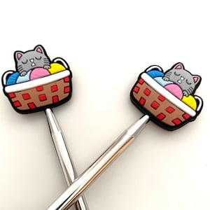 May include: Two silver knitting needles with colourful cat-themed tops. The tops feature a grey cat sleeping in a basket with balls of yarn in blue, pink, yellow, and white.