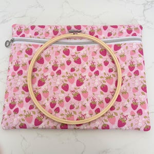May include: A pink fabric pouch with a strawberry print, featuring a silver zip and a wooden embroidery hoop. The pouch is covered in a pattern of pink strawberries with green leaves and gold details. The embroidery hoop is made of light wood.