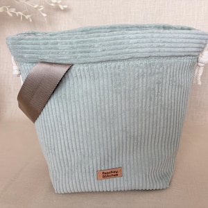 May include: A light sage green corduroy bucket bag with a drawstring closure featuring white rope ties. The bag includes a brown nylon shoulder strap and a small rectangular leather-like patch at the bottom centre with the text Peachey Stitches.