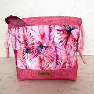 May include: A drawstring pouch featuring a white and pink tropical palm leaf print on the center panel, framed by solid pink textured fabric at the top and bottom. The bag includes pink cord drawstrings and a brown label reading Peachey Stitches.