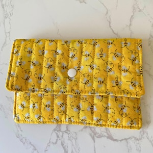 May include: A yellow quilted wallet with a bee pattern. The wallet is rectangular with a flap that closes with a white button. The bee pattern features silver and black bees on a yellow background.