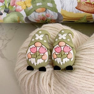 May include: Two gnome-shaped ornaments with olive green hats decorated with white stars and butterfly designs. Each gnome holds pink tulips and has a white beard, peach-coloured arms, and black shoes. The ornaments rest on a cream-coloured, textured surface.
