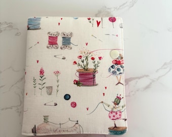 Op de afbeelding: Een notitieboek met een stoffen omslag met een naai-thema print. Het ontwerp bevat garenklosjes, bloemen, naaimachines en een knoopsluiting. De achtergrond is crèmekleurig met rode hartaccenten.