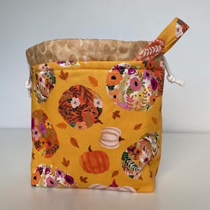 May include: A yellow drawstring bag with a floral and pumpkin print. The bag has a white drawstring and a brown lining.