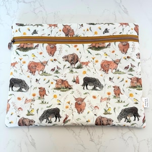 May include: A white zipped pouch featuring a repeating pattern of Highland cows, bears, foxes, birds, and flowers. The pouch has a brown zip and a quilted texture. The design has a whimsical, nature-inspired theme.