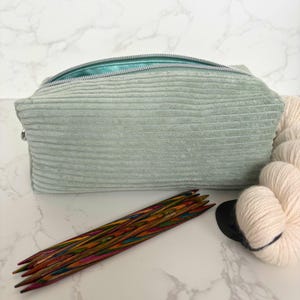 May include: A pale green corduroy zipped pouch with a teal interior, beside a bundle of cream-coloured yarn, and a set of colourful knitting needles. A black circular object is also present.