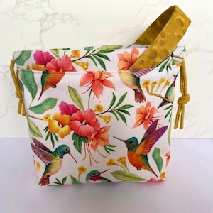 May include: A white drawstring bag with a yellow handle. The bag is decorated with a floral pattern featuring pink and yellow flowers and colorful hummingbirds.