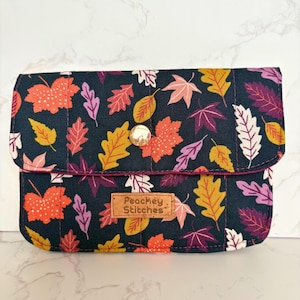 May include: A quilted clutch with a flap closure, featuring a dark navy background adorned with colorful autumn leaves in shades of orange, yellow, purple, and white. The clutch has a gold snap closure and a small leather tag that reads "Peachey Stitches."