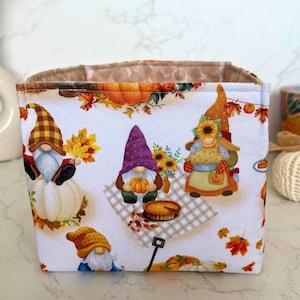 May include: Fabric storage bin with a white background featuring colorful fall gnomes, pumpkins, and autumn leaves. The gnomes are depicted with various hats and holding pumpkins. The interior of the bin is a light brown color.