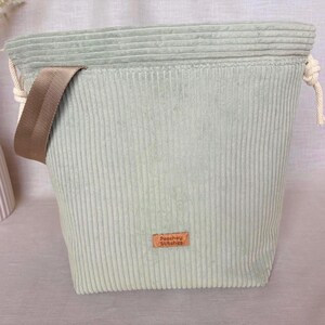 May include: A sage green corduroy drawstring bag featuring a textured ribbed surface, a wide beige woven strap attached to the side, and a small rectangular brown leather-like label centred on the front with the text Peachey Stitches printed.
