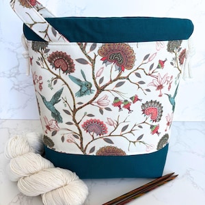 May include: A teal and white yarn storage bag with a floral and hummingbird pattern. The bag has a teal base and rim, a white handle, and a drawstring closure. A skein of white yarn and knitting needles are next to the bag.