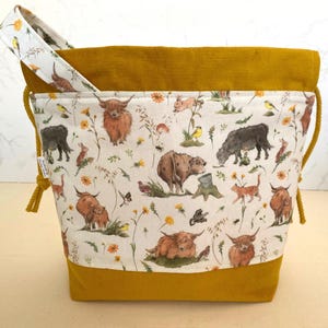 May include: A mustard yellow drawstring bag with a white fabric lining featuring a repeating pattern of Highland cows, birds, and flowers. The bag has a mustard yellow corduroy top and bottom.