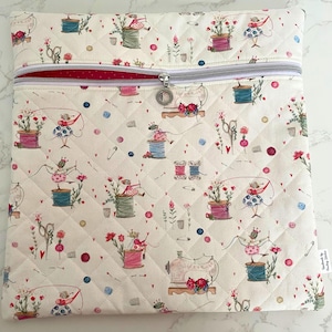 May include: Cream quilted fabric bag with a zip closure. The bag features a repeating pattern of sewing-themed illustrations, including spools of thread, sewing machines, and figures. The interior lining is red. The zip pull has a small charm.