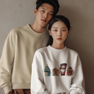 以下が含まれることがあります： カップルがお揃いのスウェットシャツを着ています。女性のトレーナーは、ジンジャーブレッドマン、サンタクロース、クリスマスツリーが描かれたマグカップがデザインされた白いクリスマスモチーフです。男性のトレーナーはベージュです。