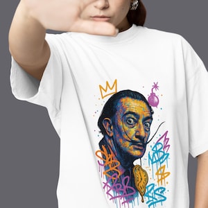 May include: White t-shirt featuring a colourful graphic of Salvador Dali with a crown, a dripping clock, and graffiti-style lettering.