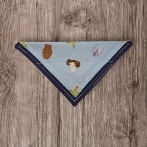 May include: A light blue, triangular pet bandana with a navy blue border. The bandana features a repeating pattern of cartoon characters, including Chewbacca, Princess Leia, Yoda, and R2-D2. The bandana is made of fabric and is likely for a dog or cat.