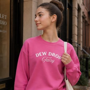 May include: A bright pink long-sleeve crewneck sweatshirt featuring the white text DEW DROP Fairy printed on the chest. The sweatshirt is paired with a light pink wrap-style skirt and a plain off-white canvas tote bag carried over the shoulder.