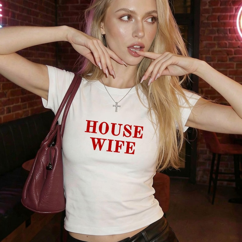 Puede incluir: Una camiseta blanca de manga corta con el texto HOUSE WIFE impreso en letras may&uacute;sculas rojas en el pecho, acompa&ntilde;ada de un bolso de cuero color burdeos oscuro y un collar con colgante de cruz plateada en una cadena fina.