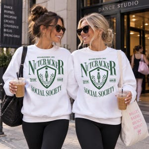May include: A white crewneck sweatshirt featuring a dark green graphic print that reads "ESTABLISHED 1892 NUTCRACKER SOCIAL SOCIETY" with a central shield emblem containing a nutcracker figure, laurel branches, and the words "CAFFEINE" and "GASOLINE".