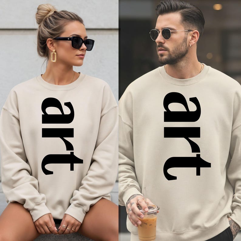 May include: Two people wearing matching beige sweatshirts with the word "art" printed vertically in large, black letters. The sweatshirts are long-sleeved and appear to be made of a soft material. The image suggests a casual, artistic theme.