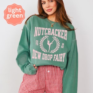 May include: A light green sweatshirt with the text "NUTCRACKER" above a ballerina graphic and "DEW DROP FAIRY" below. The sweatshirt has long sleeves and a crew neck. The model is wearing red and white checkered shorts.