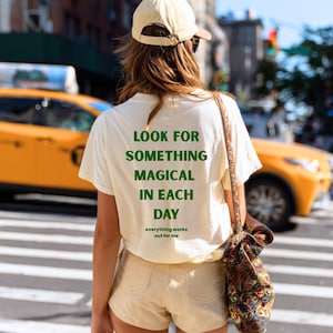 Peut inclure: Un t-shirt crème avec le texte "LOOK FOR SOMETHING MAGICAL IN EACH DAY" en vert. La phrase "everything works out for me" est imprimée en dessous. La personne porte un short beige et une casquette de baseball beige.